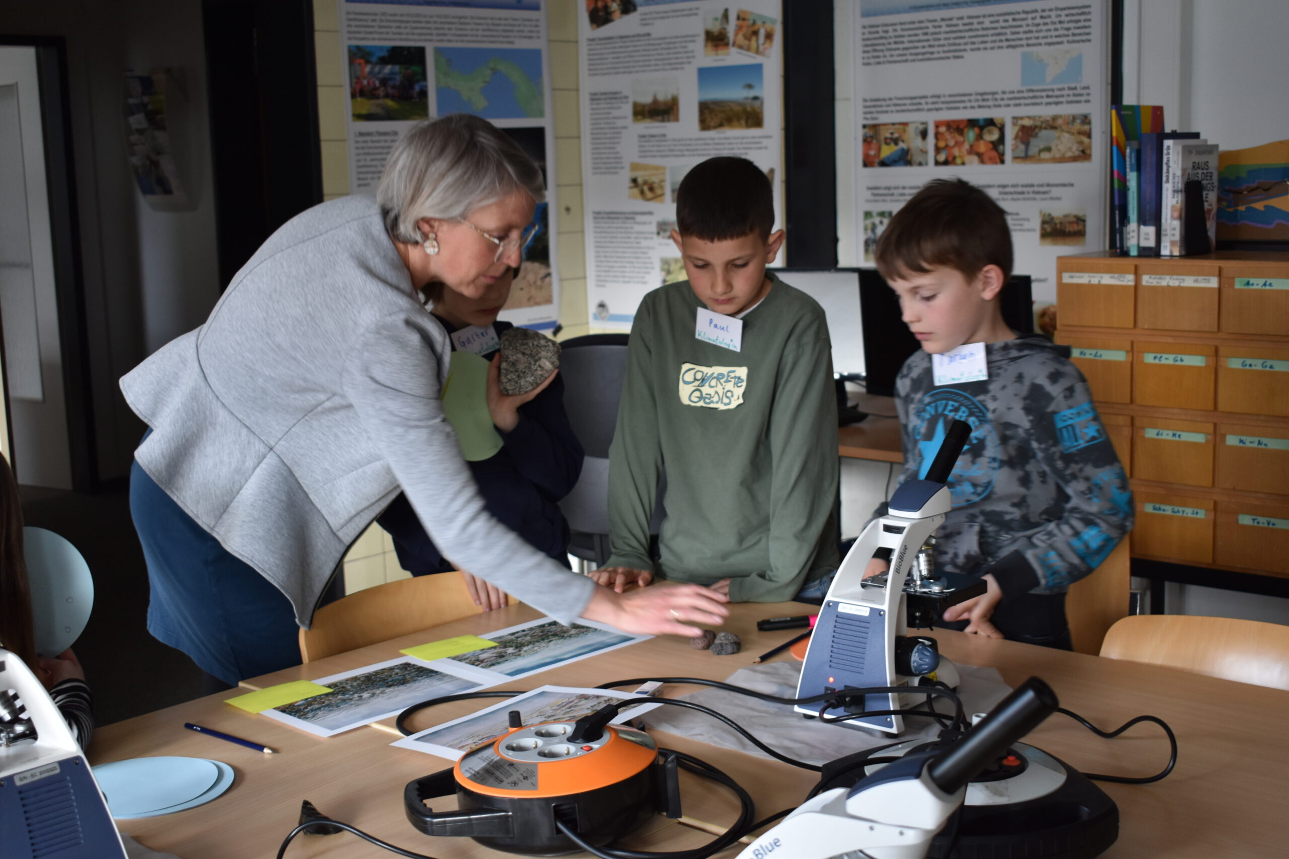 A group of children gathered around microscopes on a library table, examining specimens while a female scientist explains the climate-research station.
