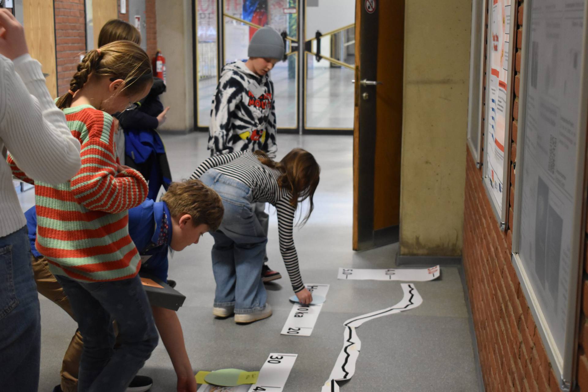 “Several children kneel and bend over in a school corridor, arranging numbered timeline strips and puzzle-piece elements on the floor to map out past climate periods.