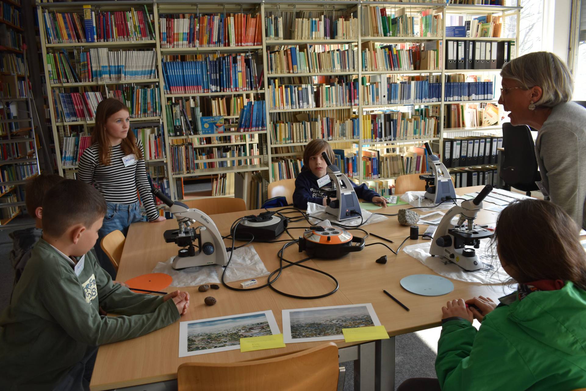 A group of children gathered around microscopes on a library table, examining specimens while a female scientist explains the climate-research station.